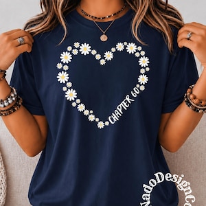 May include: A navy blue short-sleeved t-shirt featuring a heart shape composed of white daisy flowers with yellow centers. The text "CHAPTER 60" is printed in white in a cursive font along the lower right side of the heart. The logo "NadoDesignCo" is visible.