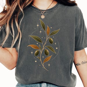 May include: A charcoal gray short-sleeve t-shirt featuring a central graphic of an olive branch with green and bronze leaves, three green olives, and small gold star accents arranged in a circular pattern on the front of the garment.