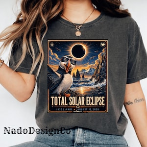 Pode incluir: Uma t-shirt cinzento-carvão com um gráfico de um papagaio-do-mar a usar óculos de sol numa praia na Islândia durante um eclipse solar total. O texto diz Total Solar Eclipse, Iceland, August 12, 2026, com uma ilustração de sol e lua acima.