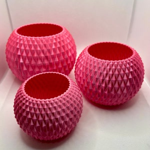 May include: Three pink, spherical planters with a geometric diamond pattern. The planters are different sizes, with a larger one in the back and a smaller one in the front. The interiors are a darker shade of pink.
