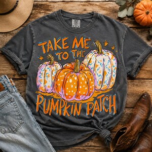 May include: A charcoal gray short-sleeve t-shirt featuring a graphic of three pumpkins with orange and blue patterns. The text reads "TAKE ME TO THE PUMPKIN PATCH" in orange letters. The shirt has a crew neckline and a vintage-style washed finish.