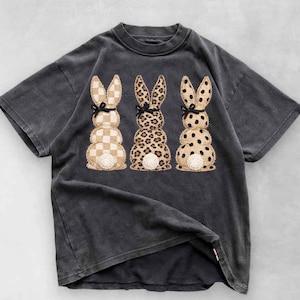 May include: Dark gray t-shirt featuring three stacked bunny designs. The bunnies have different patterns: checkered, leopard print, and polka dots. Each bunny has a black bow. The shirt has a vintage, washed look.