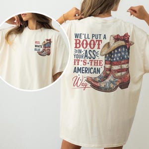 May include: Cream-colored t-shirt featuring a patriotic design. The front has a small graphic of a cowboy boot with the text "RED, WHITE, BLUE." The back displays a larger graphic of a cowboy boot with the American flag and the text "WE'LL PUT A BOOT IN YOUR ASS IT'S THE AMERICAN WAY."