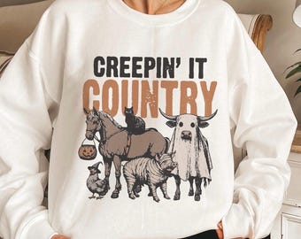 May include: White sweatshirt with the text "CREEPIN' IT COUNTRY" in black and brown. The graphic features a horse, cat, cow, pig, and chicken, all dressed in Halloween costumes. A pumpkin bucket hangs from the horse.