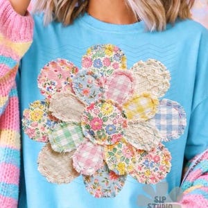 May include: A light blue t-shirt featuring a large floral design made of various fabric patches with floral and gingham patterns. The sleeves are covered by a pastel striped sweater. The text "SIP STUDIO" is visible at the bottom.