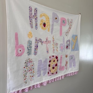 Könnte beinhalten: Ein weißer Wandbehang aus Stoff mit dem applizierten Text Happy Birthday AMELIA aus verschiedenen bunten Stoffen mit Blumen-, Vichy- und Erdbeermustern. Das Banner ist am unteren Rand mit einer rosa Ösen-Spitzenborte versehen.