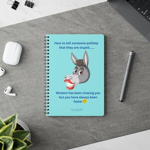 Pode incluir: Um caderno espiral com uma capa azul clara. A capa apresenta um burro de desenho animado e o texto "How to tell someone politely that they are stupid..." e "Wisdom has been chasing you but you have always been faster."