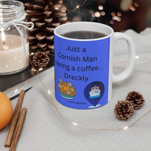 May include: A blue ceramic mug with a white handle. The mug has a white rim and a white interior. The mug has a graphic of a Cornish pasty, a boat, and the text "Just a Cornish Man having a coffee... Dreckly". The text "InspiringByKate" is at the bottom of the mug.