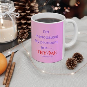 Op de afbeelding: Een roze koffiemok met de tekst "I'm menopausal. My pronouns are... TRY/ME" in paarse en rode tekst. De mok staat op een wit oppervlak met een bruine dennenappel en een glazen pot.