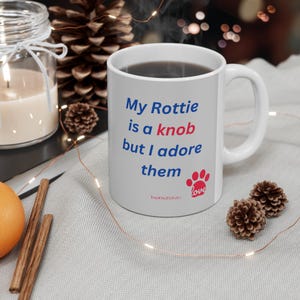 May include: White ceramic coffee mug with a blue and red graphic that says "My Rottie is a knob but I adore them" with a paw print and the word "love" in red.