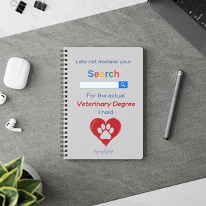 Pode incluir: Um caderno espiral cinzento com o texto "Lets not mistake your Search For the actual Veterinary Degree I hold." O design inclui uma barra de pesquisa e um coração vermelho com uma pegada.