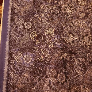 May include: A dark blue textile with a complex floral pattern in white and lighter blue. The fabric features a detailed design of flowers and leaves, with a solid blue border along one edge. The overall effect is intricate and decorative.