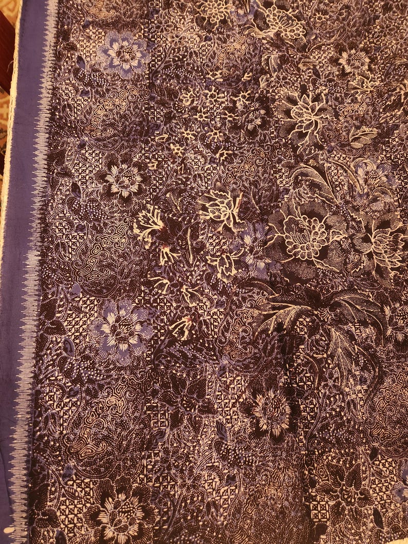 May include: A dark blue textile with a complex floral pattern in white and lighter blue. The fabric features a detailed design of flowers and leaves, with a solid blue border along one edge. The overall effect is intricate and decorative.