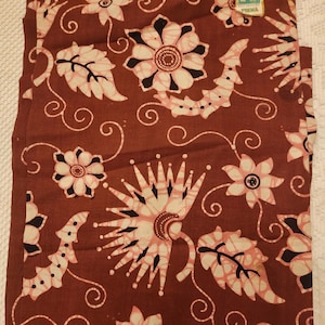 May include: A folded brown fabric with a floral pattern in white and pink. The design includes stylized flowers, leaves, and swirling lines. A small tag in the upper right corner reads "Batik Semar Prima".