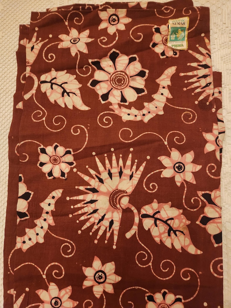 May include: A folded brown fabric with a floral pattern in white and pink. The design includes stylized flowers, leaves, and swirling lines. A small tag in the upper right corner reads "Batik Semar Prima".