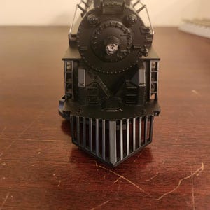 May include: A detailed, black model train engine, viewed head-on. The engine features a prominent headlight, a circular front, and a slatted cowcatcher. The model sits on a wooden surface, showcasing its intricate design and classic appeal.