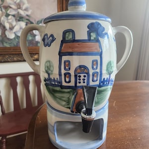 Puede incluir: Un dispensador de bebidas de cerámica con un motivo de casa pintado a mano en azul y blanco, con dos asas laterales, una tapa cónica con rayas azules y un grifo de plástico negro sobre una base cilíndrica a juego con aberturas.