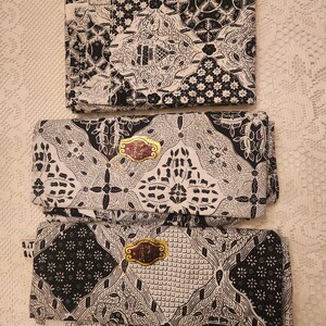 May include: Three folded batik fabric pieces with intricate black and white patterns. The designs include floral and geometric motifs. Each piece has a gold-colored label. The fabric is displayed on a white, textured surface.