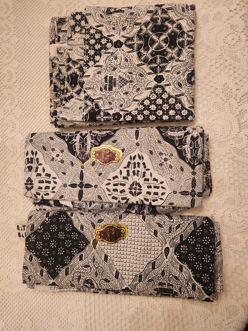 May include: Three folded batik fabric pieces with intricate black and white patterns. The designs include floral and geometric motifs. Each piece has a gold-colored label. The fabric is displayed on a white, textured surface.