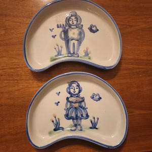 Puede incluir: Dos platos de cerámica blancos en forma de media luna con borde azul. Cada plato presenta una ilustración pintada a mano de una persona con atuendo azul, rodeada de plantas y nubes estilizadas. Los platos están sobre una superficie de madera.