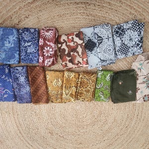 May include: An assortment of folded fabric pieces with various patterns and colors. The textiles include floral, geometric, and abstract designs in shades of blue, brown, gold, green, and white. The fabrics are arranged on a textured, light brown surface.