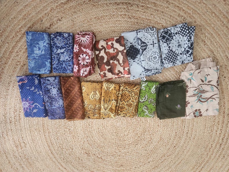 May include: An assortment of folded fabric pieces with various patterns and colors. The textiles include floral, geometric, and abstract designs in shades of blue, brown, gold, green, and white. The fabrics are arranged on a textured, light brown surface.