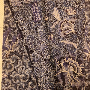 May include: A textile with a dark blue background and a white floral pattern. The design includes large, stylized flowers and intricate leaf and swirl details. The fabric has a textured appearance, with a label that reads "BAIK HALUS SINAR KENCANA".