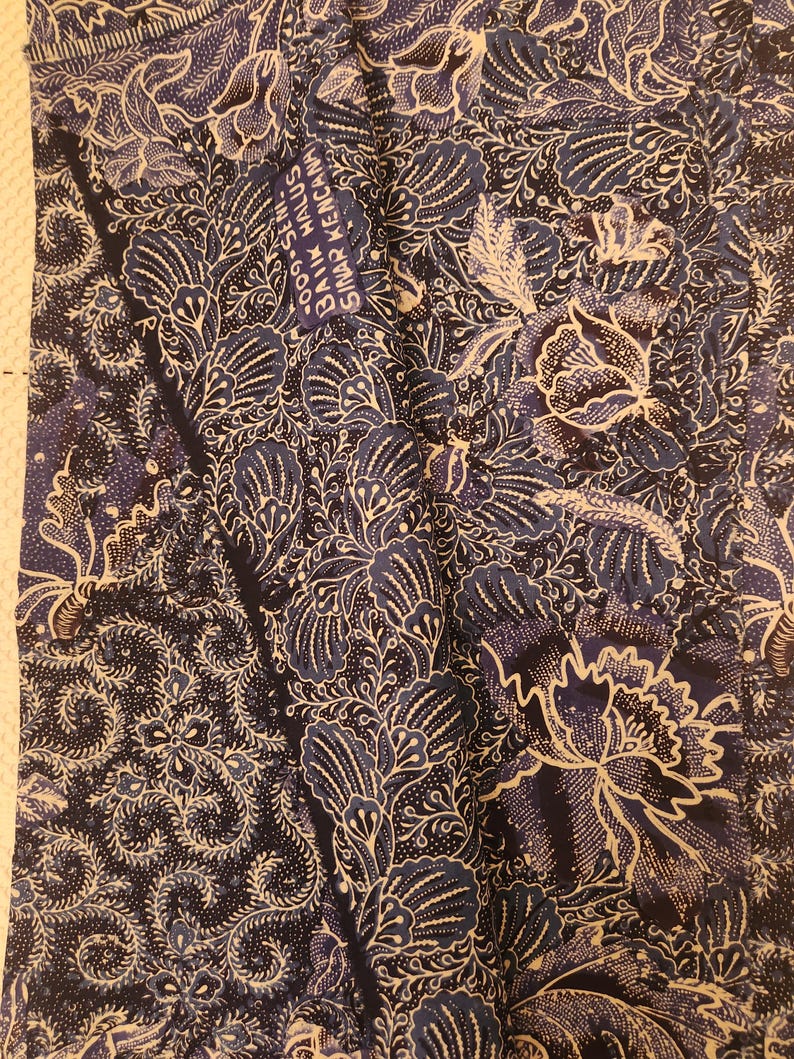 May include: A textile with a dark blue background and a white floral pattern. The design includes large, stylized flowers and intricate leaf and swirl details. The fabric has a textured appearance, with a label that reads "BAIK HALUS SINAR KENCANA".