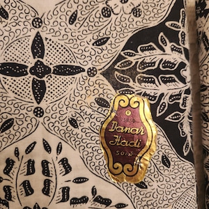 May include: Close-up of a fabric with a detailed black and white batik pattern. The design includes floral and geometric shapes. A gold and burgundy label reads "Danar Hadi SOLO".