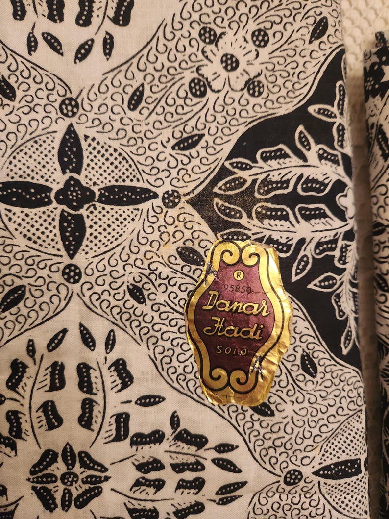 May include: Close-up of a fabric with a detailed black and white batik pattern. The design includes floral and geometric shapes. A gold and burgundy label reads "Danar Hadi SOLO".