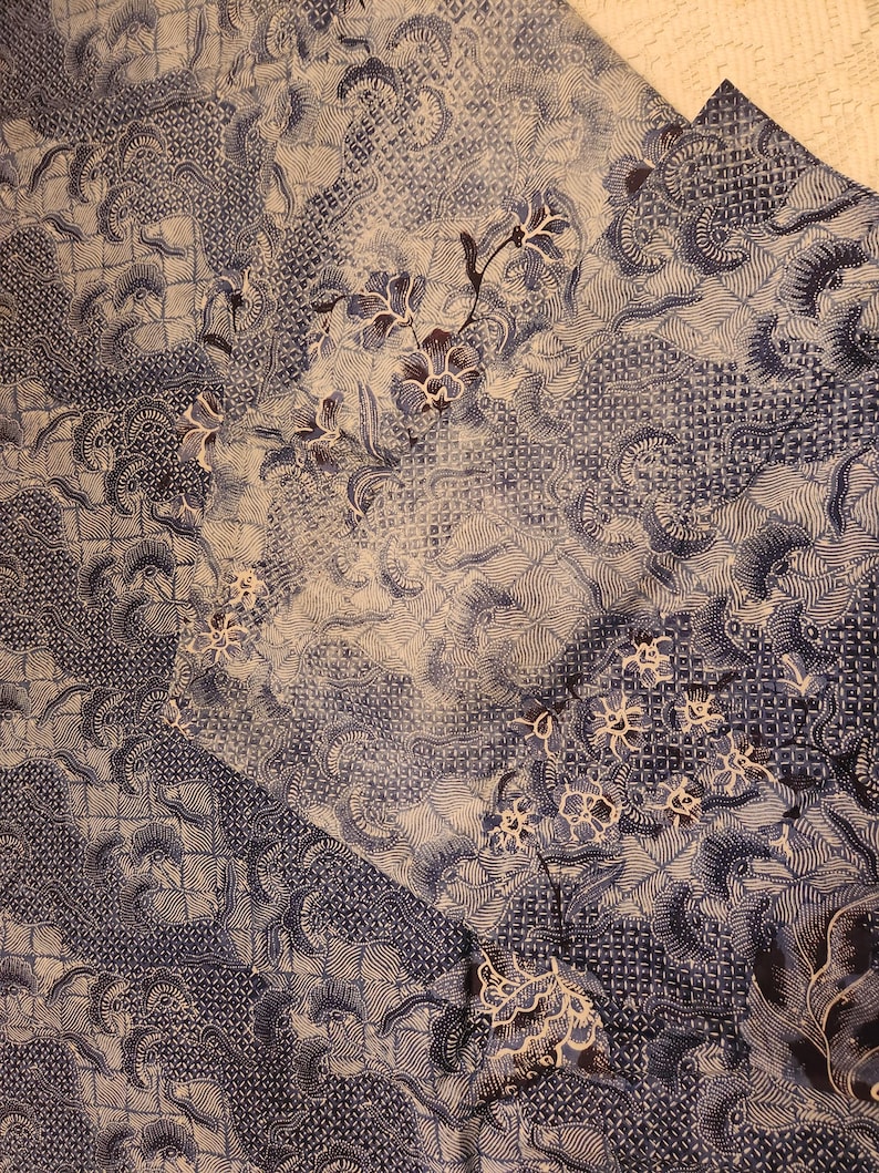 May include: Close-up of a blue and white patterned fabric with floral and geometric designs. The fabric has a batik-style appearance, with intricate details and a textured surface. The color palette is primarily shades of blue and white.