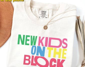 May include: Ivory-colored t-shirt with the text "NEW KIDS ON THE BLOCK" in green, pink, blue, and yellow. A gold necklace with a pendant rests on the shirt. The shirt has a Comfort Colors tag.