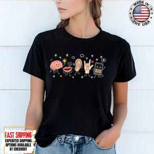 May include: A black short-sleeve t-shirt featuring a graphic print of cartoon characters including a brain, lips, an ear, a hand making a sign language gesture, and a smartphone, all surrounded by small colorful stars, hearts, and speech bubbles.