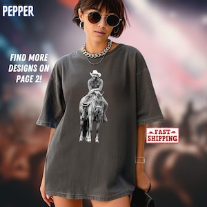 May include: A charcoal grey short-sleeve t-shirt featuring a black and white graphic print of a cowboy riding a horse. The shirt has a crew neckline and a relaxed fit. The text PEPPER, FIND MORE DESIGNS ON PAGE 2!, and FAST SHIPPING are visible.