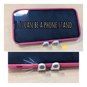 May include: A pink phone case with white paw print phone stand attachments. The phone case is on a wooden surface. The text "IT CAN BE A PHONE STAND" is visible on the phone screen.