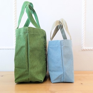 May include: Two tote bags, one green and one light blue, standing side by side on a wooden surface. The green bag has Japanese text on the side. Both bags have a simple design with two handles.