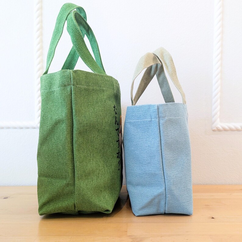 May include: Two tote bags, one green and one light blue, standing side by side on a wooden surface. The green bag has Japanese text on the side. Both bags have a simple design with two handles.