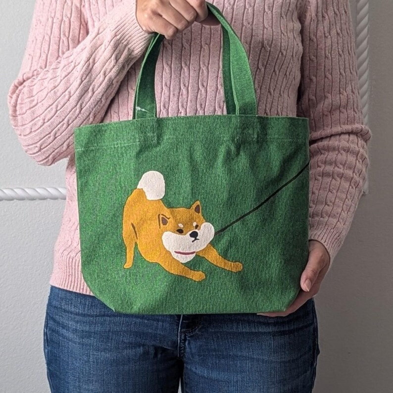 May include: A green canvas tote bag with a brown and white Shiba Inu dog illustration. The dog is pulling on a black leash and has a playful expression.