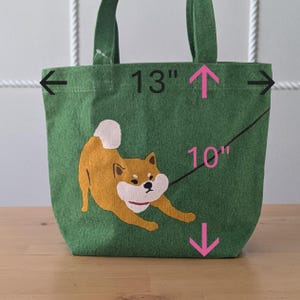 May include: Green canvas tote bag with a Shiba Inu dog graphic. The bag measures 13 inches wide and 10 inches tall.