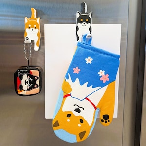 May include: A set of three oven mitts with a Shiba Inu dog design. The mitts are blue, white, and orange with a floral pattern. The mitts are hanging on a refrigerator with a magnet.