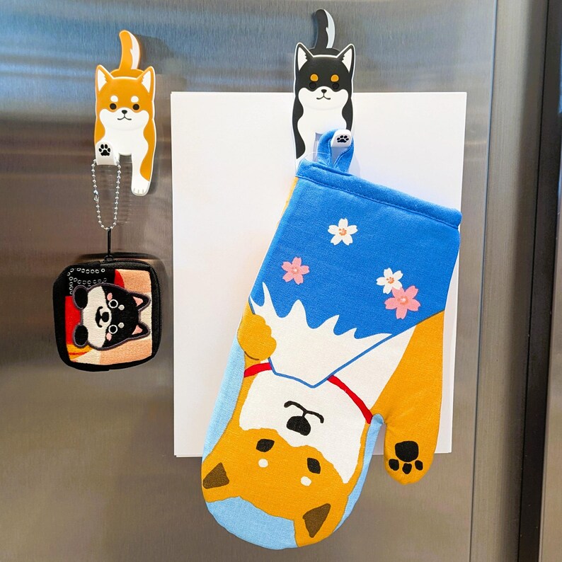 May include: A set of three oven mitts with a Shiba Inu dog design. The mitts are blue, white, and orange with a floral pattern. The mitts are hanging on a refrigerator with a magnet.