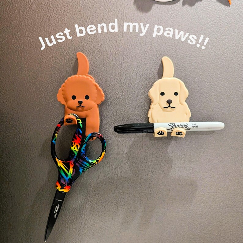 May include: Two dog-shaped magnetic hooks on a grey surface. The brown dog hook holds a pair of rainbow-colored scissors. The beige dog hook holds a black marker with the text "Sharpie Fine Point" on it. The text "Just bend my paws!" is written above the hooks.
