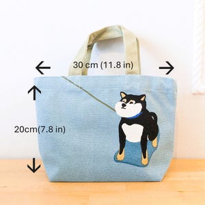 May include: A light blue tote bag with a black and white Shiba Inu dog illustration. The bag has a light brown canvas handle and measures 11.8 inches wide and 7.8 inches tall.