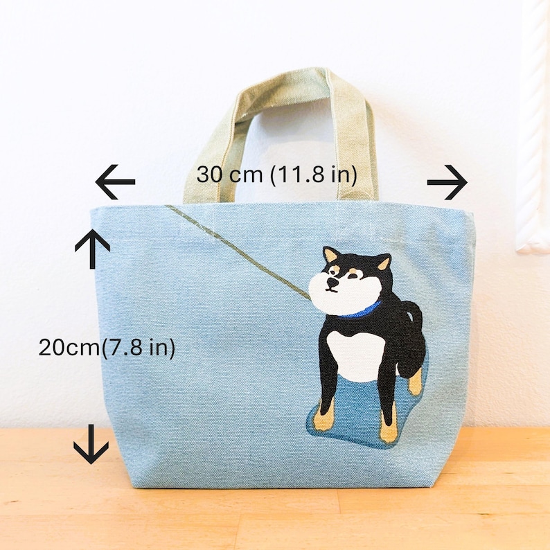 May include: A light blue tote bag with a black and white Shiba Inu dog illustration. The bag has a light brown canvas handle and measures 11.8 inches wide and 7.8 inches tall.