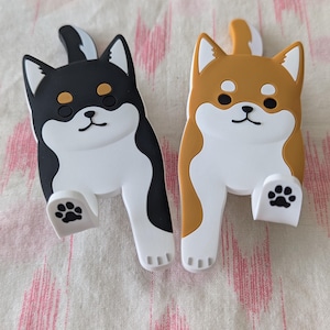 May include: Two adorable Shiba Inu dog-shaped phone holders. One is black and white, the other is brown and white.