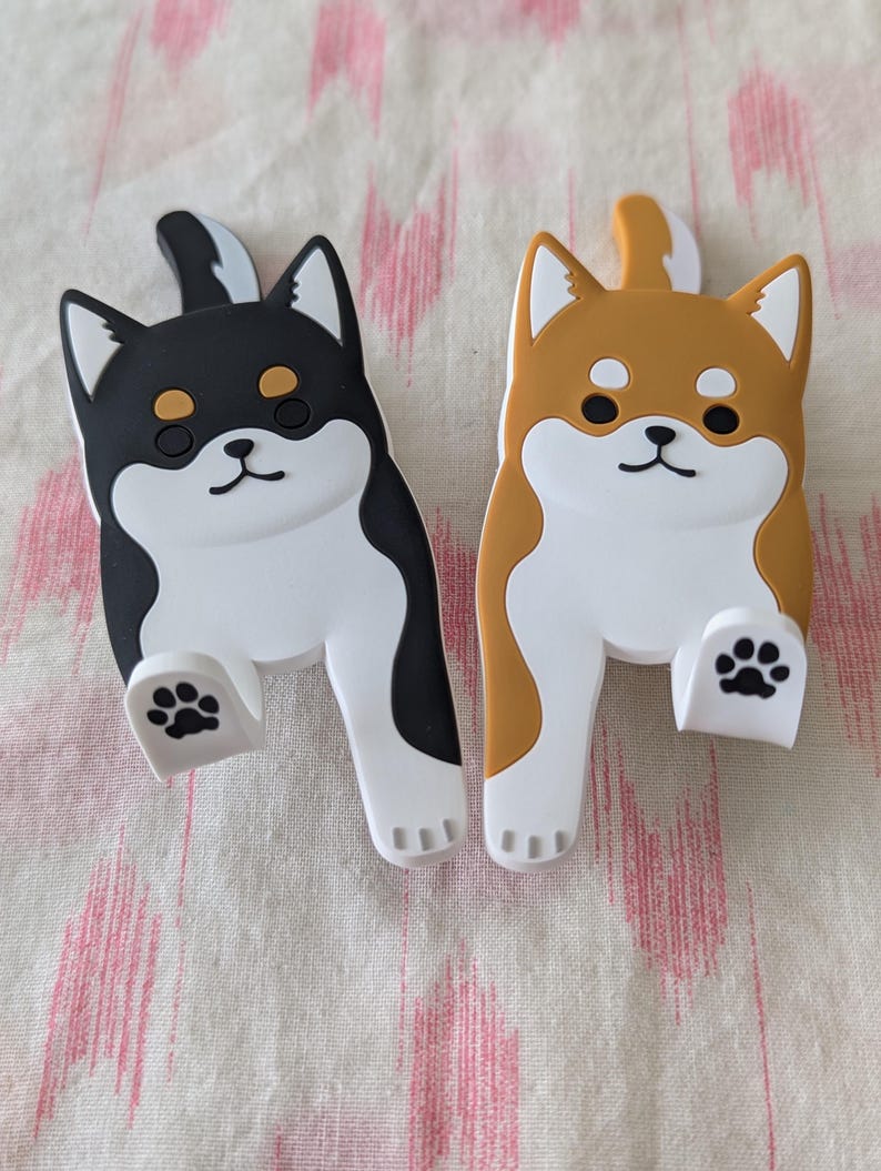 May include: Two adorable Shiba Inu dog-shaped phone holders. One is black and white, the other is brown and white.