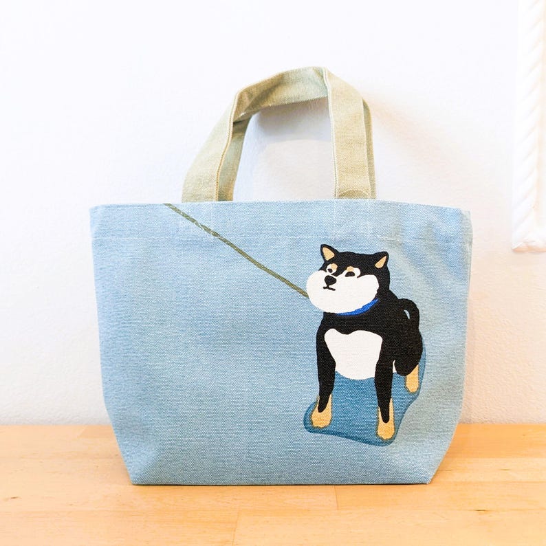 May include: A light blue canvas tote bag with a black and white Shiba Inu dog illustration. The dog is wearing a blue collar and is on a leash. The bag has two beige canvas handles.