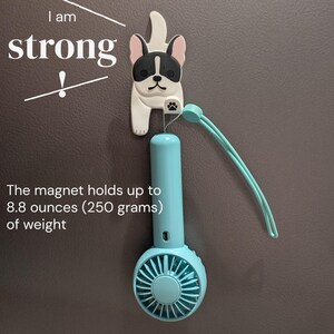 May include: A small, blue, handheld fan with a white, plastic, bulldog-shaped magnet attached to the top. The magnet holds up to 8.8 ounces (250 grams) of weight. The text "I am strong!" is written on the wall behind the fan.