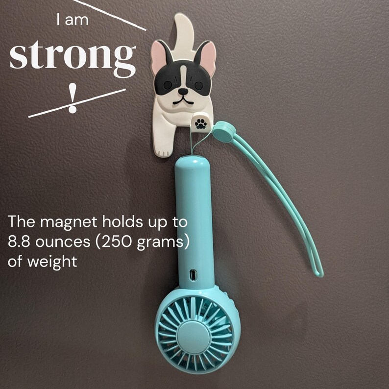 May include: A small, blue, handheld fan with a white, plastic, bulldog-shaped magnet attached to the top. The magnet holds up to 8.8 ounces (250 grams) of weight. The text "I am strong!" is written on the wall behind the fan.