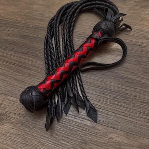 May include: A black leather whip with a red and black woven handle. The whip has a loop at the end of the handle and multiple black leather strands. The handle is approximately 6 inches long.